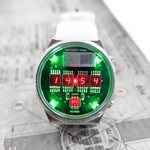 May include: A silver wristwatch with a green and red digital display. The watch face features a circuit board design with the text "NIXOID" and "AM/PM". The time is displayed as "1:45:4".