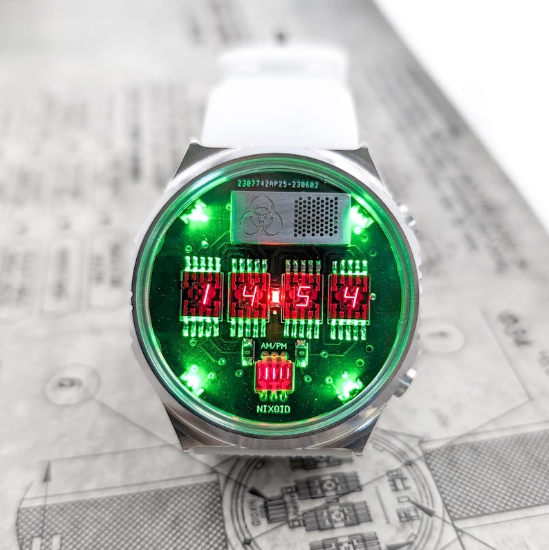 May include: A silver wristwatch with a green and red digital display. The watch face features a circuit board design with the text "NIXOID" and "AM/PM". The time is displayed as "1:45:4".