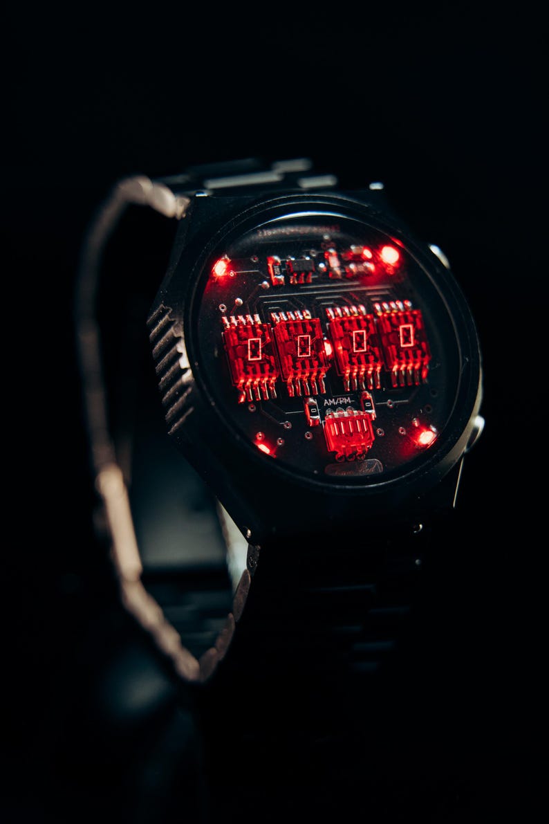 May include: Black wristwatch with a unique circuit board design. The watch face features glowing red lights and a digital display. The watchband is black and metallic. The overall aesthetic is futuristic and tech-inspired.