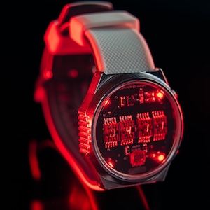 May include: A futuristic digital wristwatch with a silver case and a gray strap. The watch face displays red illuminated numbers and circuit board details. The time displayed is 04:17. The watch is set against a black background.