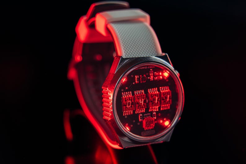 May include: A futuristic digital wristwatch with a silver case and a gray strap. The watch face displays red illuminated numbers and circuit board details. The time displayed is 04:17. The watch is set against a black background.