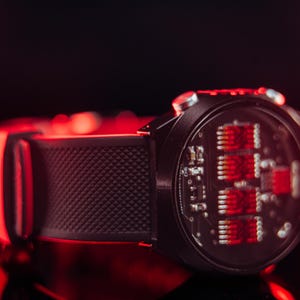 May include: A black smartwatch with a textured band and a red-lit circuit board display. The watch is set against a dark background with red highlights, showcasing its modern design and technology.