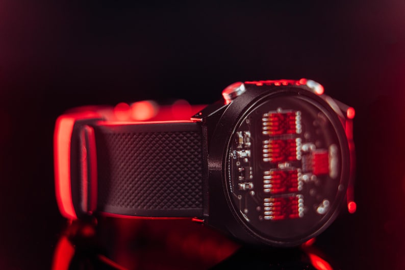 May include: A black smartwatch with a textured band and a red-lit circuit board display. The watch is set against a dark background with red highlights, showcasing its modern design and technology.