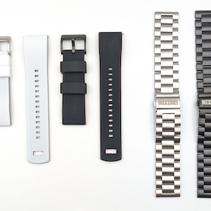 May include: Five different watch bands, including a white silicone band, a black silicone band, a black rubber band, a silver metal band, and a black metal band. The metal bands have the word "NIXON" engraved on the clasp.