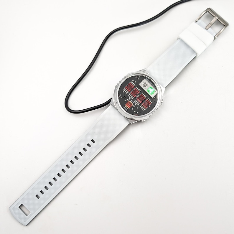May include: White digital watch with a black face and red and green digital display. The watch is connected to a black charging cable.