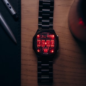 May include: A black wristwatch with a digital display showing red numerals. The watch has a black metal band and a round face. The watch is on a wooden surface, with a pen and a notebook in the background.