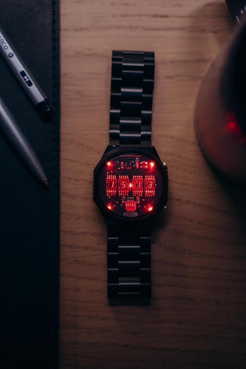 May include: A black wristwatch with a digital display showing red numerals. The watch has a black metal band and a round face. The watch is on a wooden surface, with a pen and a notebook in the background.