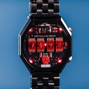 Può includere: Orologio digitale nero con quadrante ottagonale e cinturino in metallo abbinato. Il quadrante mostra l'ora in numeri rossi, con dettagli del circuito stampato. Lo sfondo è blu.