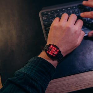 May include: A black digital watch with a red LED display is worn on a wrist. The watch has a rectangular face and a metal band. The person is wearing a green and black plaid shirt and typing on a keyboard.