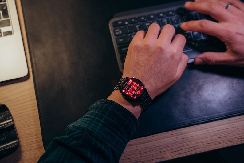 May include: A black digital watch with a red LED display is worn on a wrist. The watch has a rectangular face and a metal band. The person is wearing a green and black plaid shirt and typing on a keyboard.