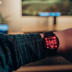 May include: Black digital watch with a red illuminated display. The watch is on a person's wrist, with a dark green and black plaid shirt sleeve visible. The watch face displays a complex circuit board design.