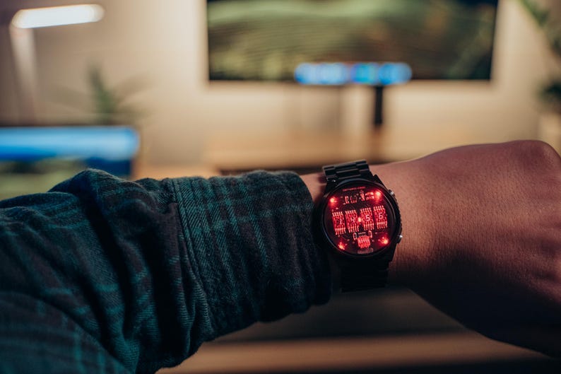 May include: Black digital watch with a red illuminated display. The watch is on a person's wrist, with a dark green and black plaid shirt sleeve visible. The watch face displays a complex circuit board design.