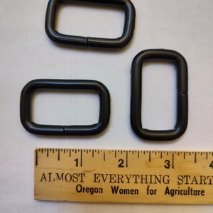 May include: Three black metal D-rings of different sizes. The rings are shown on a white surface with a wooden ruler marked with inches.