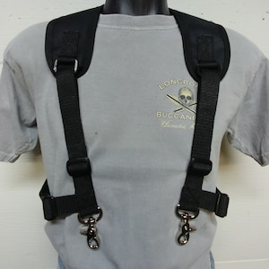 May include: Black adjustable camera harness with two metal clips. The harness is worn over a grey t-shirt with the text "Longboard Buccane" and "Hanaiki, Hawaii" printed on it.