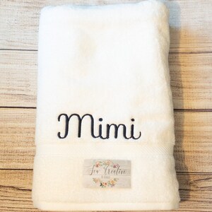Puede incluir: Toalla de ba&ntilde;o blanca con el nombre "Mimi" bordado en letras cursivas negras. La toalla tiene una peque&ntilde;a etiqueta con el texto "Sew Creative by Kelli" en letras negras.