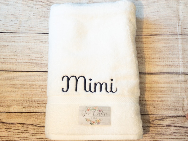 Puede incluir: Toalla de ba&ntilde;o blanca con el nombre "Mimi" bordado en letras cursivas negras. La toalla tiene una peque&ntilde;a etiqueta con el texto "Sew Creative by Kelli" en letras negras.