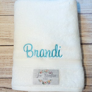 May include: White bath towel with the name "Brandi" embroidered in teal blue lettering. The towel has a small tag that says "Sew Creative by Addie".
