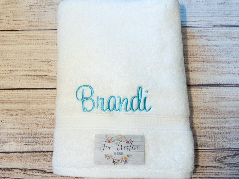 Puede incluir: Toalla de ba&ntilde;o blanca con el nombre "Brandi" bordado en letras azul turquesa. La toalla tiene una peque&ntilde;a etiqueta que dice "Sew Creative by Addie".