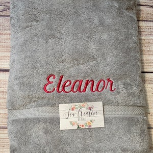 Puede incluir: Una toalla de ba&ntilde;o gris con el nombre "Eleanor" bordado en hilo rojo. La toalla tiene un borde blanco y una peque&ntilde;a etiqueta blanca con el texto "Sew Creative by Ashlee".