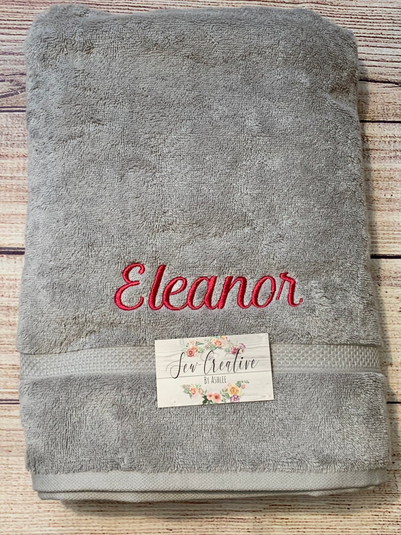 Puede incluir: Una toalla de ba&ntilde;o gris con el nombre "Eleanor" bordado en hilo rojo. La toalla tiene un borde blanco y una peque&ntilde;a etiqueta blanca con el texto "Sew Creative by Ashlee".