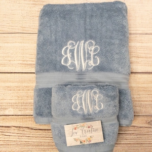 May include: A blue bath towel with a white monogram embroidered on it. The towel is folded in half and has a small tag that says "Sew Creative by Kristin".