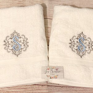 Puede incluir: Dos toallas de ba&ntilde;o blancas con un monograma bordado en azul y gris de la letra "S" en un dise&ntilde;o decorativo. Las toallas est&aacute;n dobladas y se muestran sobre una superficie de madera. El texto "Sew Creative by Kristi" est&aacute; en una peque&ntilde;a etiqueta adjunta a una de las toallas.