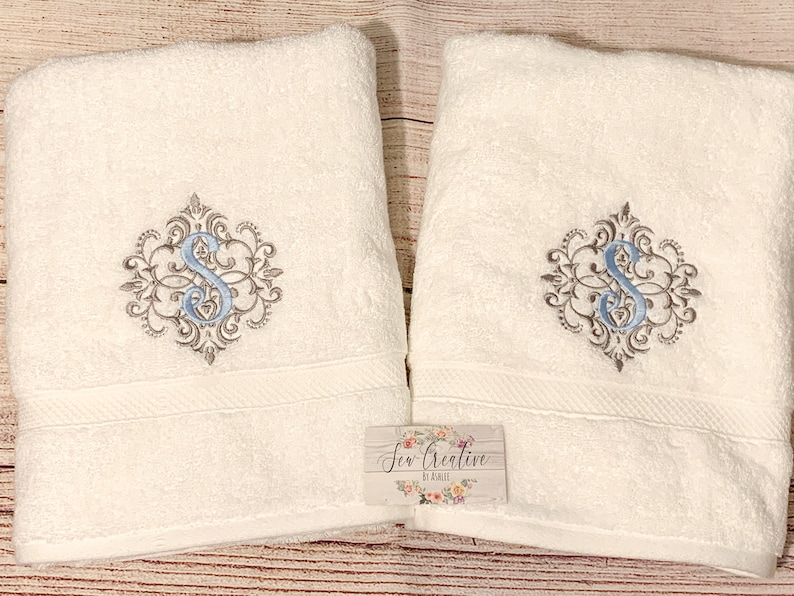Puede incluir: Dos toallas de ba&ntilde;o blancas con un monograma bordado en azul y gris de la letra "S" en un dise&ntilde;o decorativo. Las toallas est&aacute;n dobladas y se muestran sobre una superficie de madera. El texto "Sew Creative by Kristi" est&aacute; en una peque&ntilde;a etiqueta adjunta a una de las toallas.