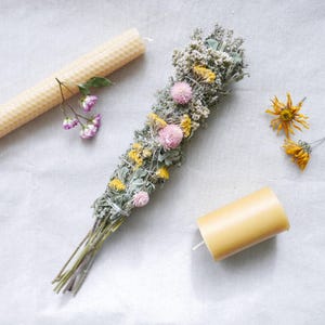 Könnte beinhalten: Draufsicht auf einen Salbei-Smudge-Stick, verziert mit rosa und gelben Trockenblumen, neben zwei Bienenwachskerzen. Eine Kerze ist lang und gerollt, die andere ist ein kurzes Zylinder. Die Artikel sind auf einer weißen Oberfläche angeordnet.