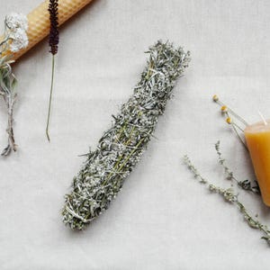 Könnte beinhalten: Ein Bündel getrockneter Salbei, mit weißer Schnur gebunden, liegt auf einer neutralfarbenen Oberfläche. Der Salbei ist eine Mischung aus Grün und Grau, mit kleinen weißen Blüten. Weitere Artikel sind eine gerollte Bienenwachskerze, getrockneter Lavendel und Zweige getrockneter Blumen.