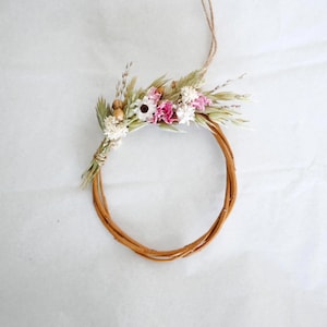 May include: A small, circular wreath made of woven brown twigs, adorned with a cluster of dried flowers in shades of white, pink, and green. A twine loop is attached for hanging. The wreath is approximately 6 inches in diameter.