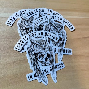 May include: A set of five black and white stickers with an owl and skull design. The stickers feature the text "Fear is not an option" in a curved font. The stickers are on a light brown wooden surface.