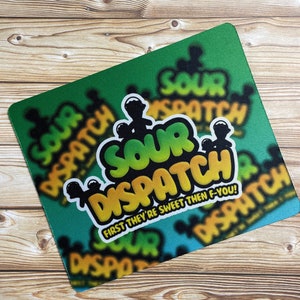 May include: A rectangular mousepad with a green and blue background. The mousepad features the words "SOUR DISPATCH" in a bold, colorful font, with the phrase "FIRST THEY'RE SWEET THEN F-YOU!" below. The design has a graffiti-style aesthetic.