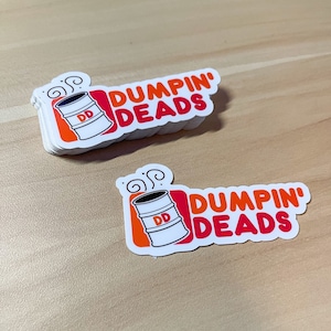 May include: A stack of white stickers with a barrel graphic and the words "DUMPIN' DEADS" in red and orange. The barrel is white with a black top, orange accents, and the letters "DD".