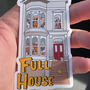 May include: A sticker featuring a gray house with multiple windows filled with cats of various colors. The sticker has the words "FULL HOUSE" in yellow block letters. A raccoon is on the roof, and cats are on the porch.
