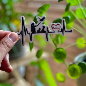 May include: A white sticker with a black heartbeat line design. The design includes silhouettes of a person, birds, and cats. A red heart with the word "Rescue" is incorporated into the design. The sticker is held against a background of green foliage.