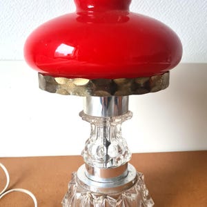 Puede incluir: Una lámpara de mesa vintage con una pantalla de cristal rojo vibrante y una base de cristal transparente facetada. La lámpara presenta detalles cromados y de latón, y un cable eléctrico blanco. La lámpara está diseñada para uso en interiores.