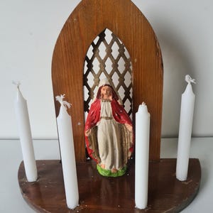Może przedstawiać: Drewniany ołtarz religijny z centralną postacią w czerwono-białej szacie, otoczoną czterema białymi świecami. Sanktuarium ma łukowaty szczyt i kratowe tło. Podstawa to zakrzywiona drewniana półka.