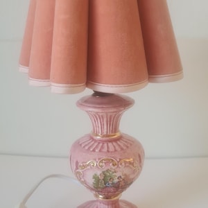 May include: A table lamp with a pleated, dusty rose fabric shade and a pink ceramic base. The base features a central oval medallion with a painted scene of figures in a landscape, accented with gold trim and a white electrical cord and plug.