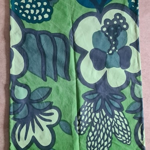 May include: Green fabric with a large floral pattern in shades of green and blue. The design features stylized flowers and leaves, creating a bold and graphic look. The fabric is likely used for home decor or apparel.
