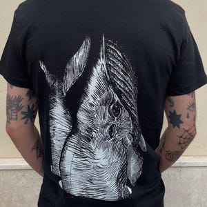 May include: Black t-shirt featuring a detailed white illustration of a creature's head on the back. The graphic has intricate linework, creating a textured appearance. The shirt is a classic crew neck style.