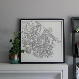 May include: A framed, white, 3D map of a city with gray lines representing streets and buildings. The map is framed in a black frame and is hanging on a white wall above a fireplace.