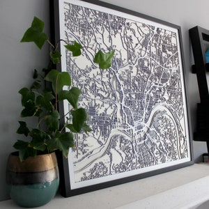 Peut inclure: Une œuvre d'art cartographique encadrée et en couches, représentant un plan de ville détaillé. La carte présente un fond blanc avec des lignes grises foncées et noires représentant les rues et les cours d'eau. L'œuvre est dans un cadre noir.