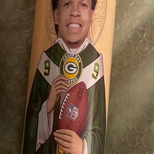 May include: A green and gold candle featuring a football player holding a football. The candle is inscribed with the words "Green Bay Packers".