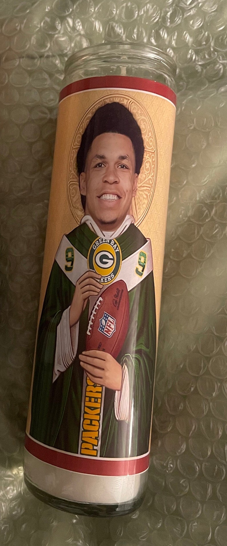 May include: A green and gold candle featuring a football player holding a football. The candle is inscribed with the words "Green Bay Packers".