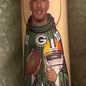 May include: A green and gold candle featuring a football player in a green and gold jersey with the number 10. The player is holding a football and wearing a helmet. The candle has a red and white border.