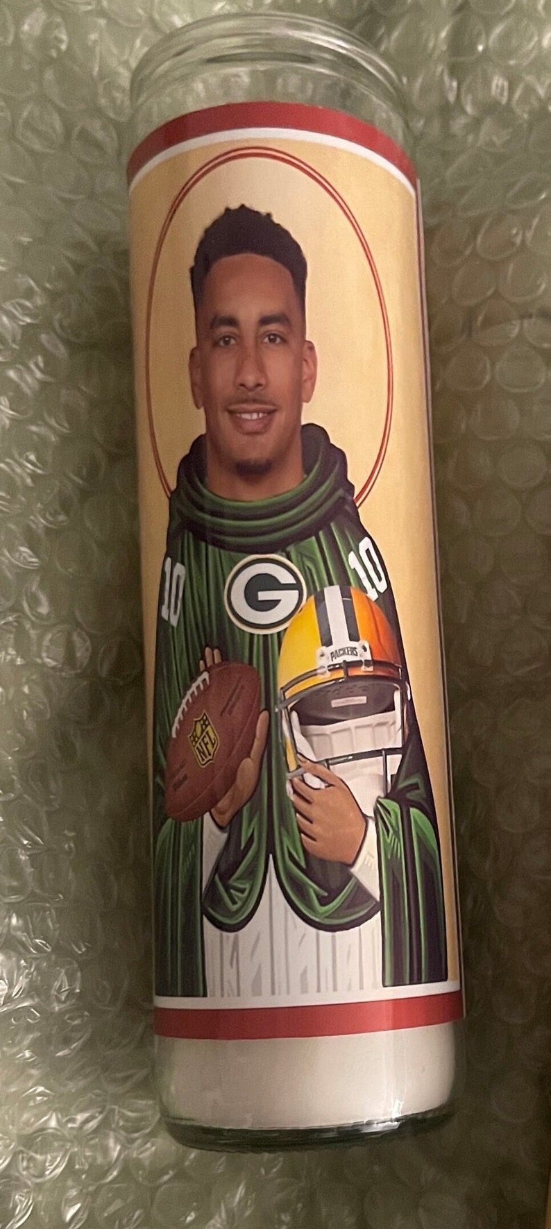 May include: A green and gold candle featuring a football player in a green and gold jersey with the number 10. The player is holding a football and wearing a helmet. The candle has a red and white border.