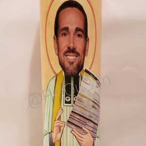 May include: A glass candle with a red, yellow and green image of a man holding a paper with text. The man is wearing a green and white striped shirt with a yellow sash.