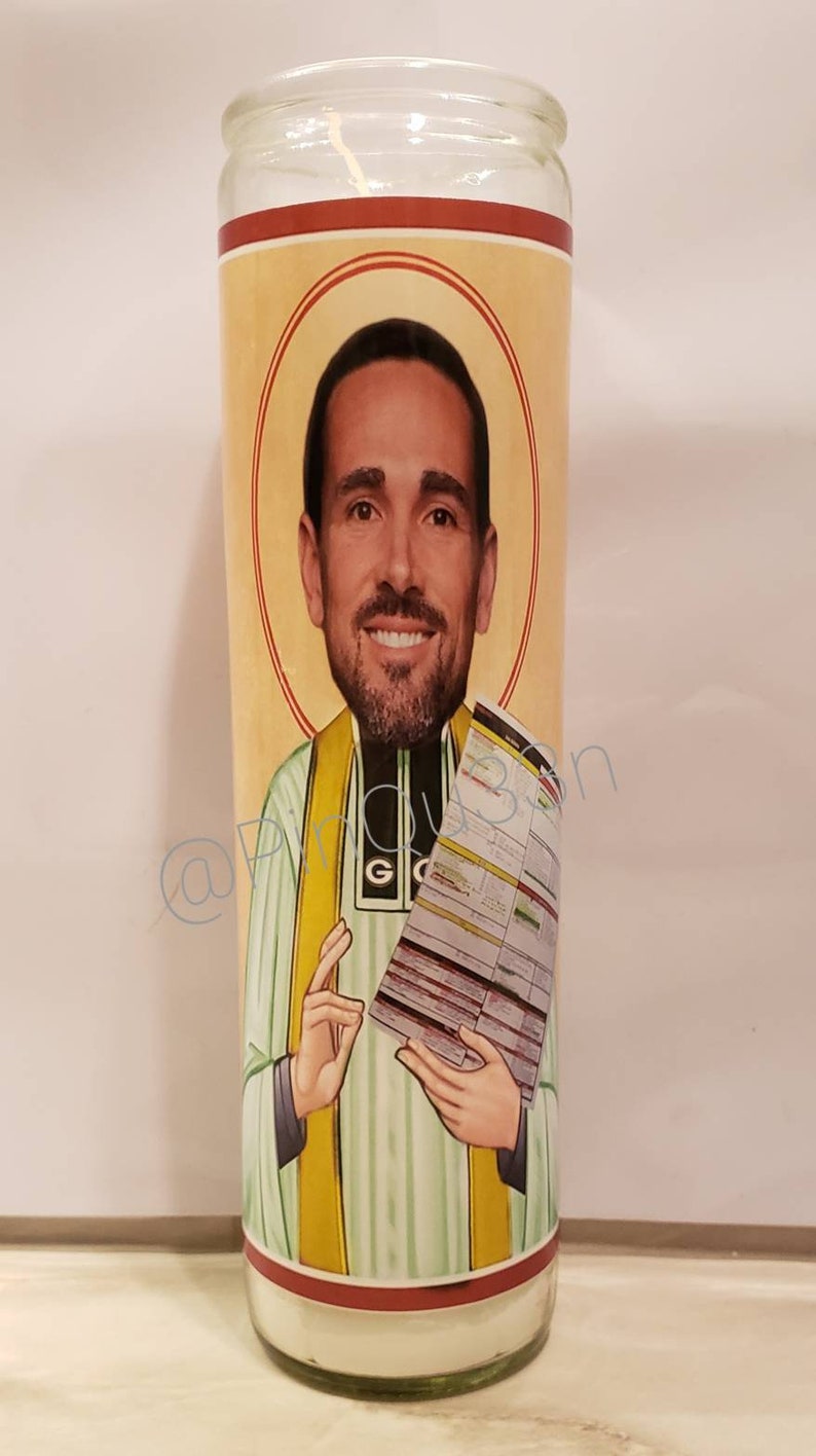 May include: A glass candle with a red, yellow and green image of a man holding a paper with text. The man is wearing a green and white striped shirt with a yellow sash.