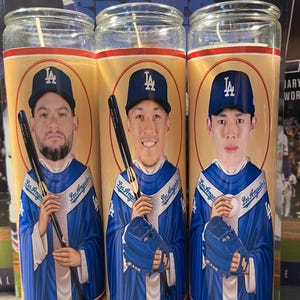 May include: Three glass prayer candles featuring Los Angeles Dodgers baseball players. Each candle has a portrait of a player wearing a blue Dodgers uniform and holding a baseball bat or glove. The candles have a yellow background with red and white accents.