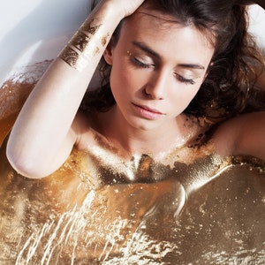 May include: A woman with long brown hair lies in a bathtub filled with gold glitter. She has gold glitter on her skin and is wearing gold glitter tattoos on her arm.